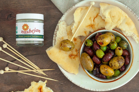 Noshejan Peak Greek Technique Marinated Olives recipe displayed on wood background with torn bread.